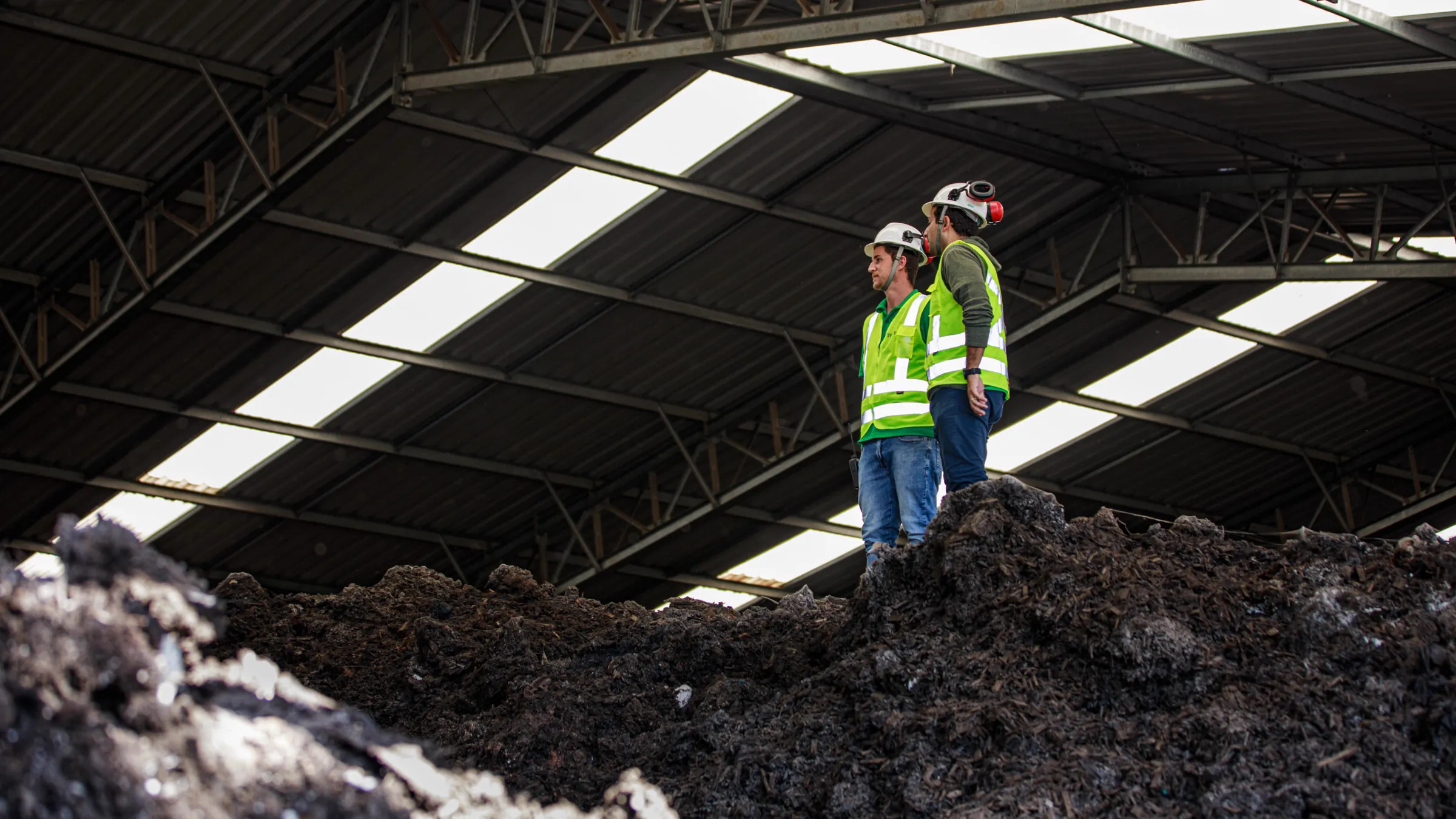Two workers in hi-vis vests and helmets standing on a compost windrow inside a covered shed
